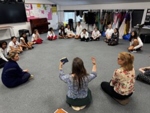 Group of students seated in a circle on the classroom floor, teacher’s hands visible as she leads the discussion in front of them