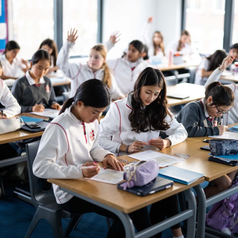 Group of students in white uniforms work at classroom desks, some raising hands and writing on papers in a bright room