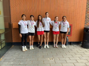 Six young athletes in white uniforms with medals pose for a photo in front of a wooden wall backdrop on a sports venue wallhanging