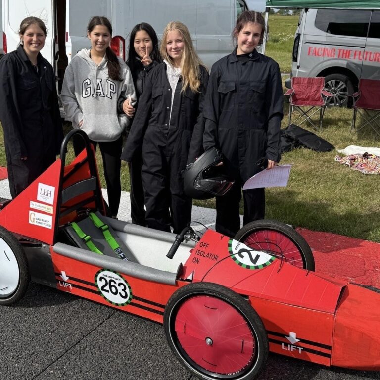 Five students in black coveralls pose with a red, open-wheel race car numbered 263 on a track; vans and tents in the background.