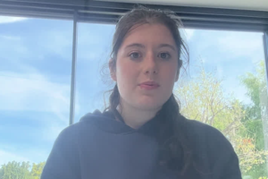 Young woman with brown hair in a loose braid wearing a black hoodie, indoors by a window with trees outside.