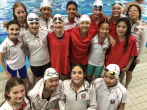 Group of smiling young swimmers in white and red uniforms posing by an indoor pool.