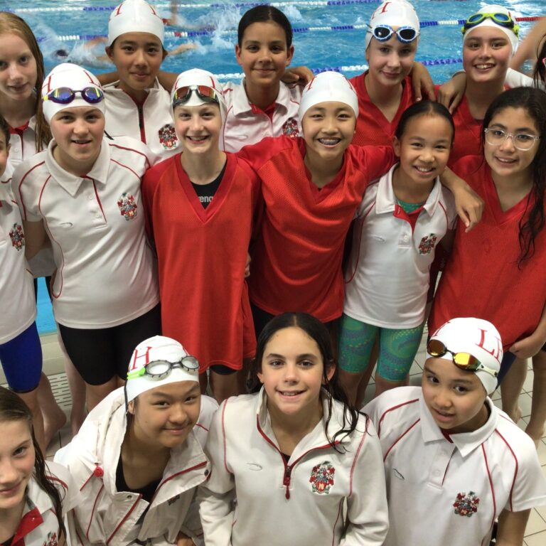 Group of smiling young swimmers in white and red uniforms posing by an indoor pool.
