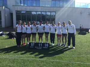 A line of young female players in white jerseys with red trim, arms around each other, standing on a grass field with a modern building behind them; scoreboard shows runs 97, overs 11, target 96.
