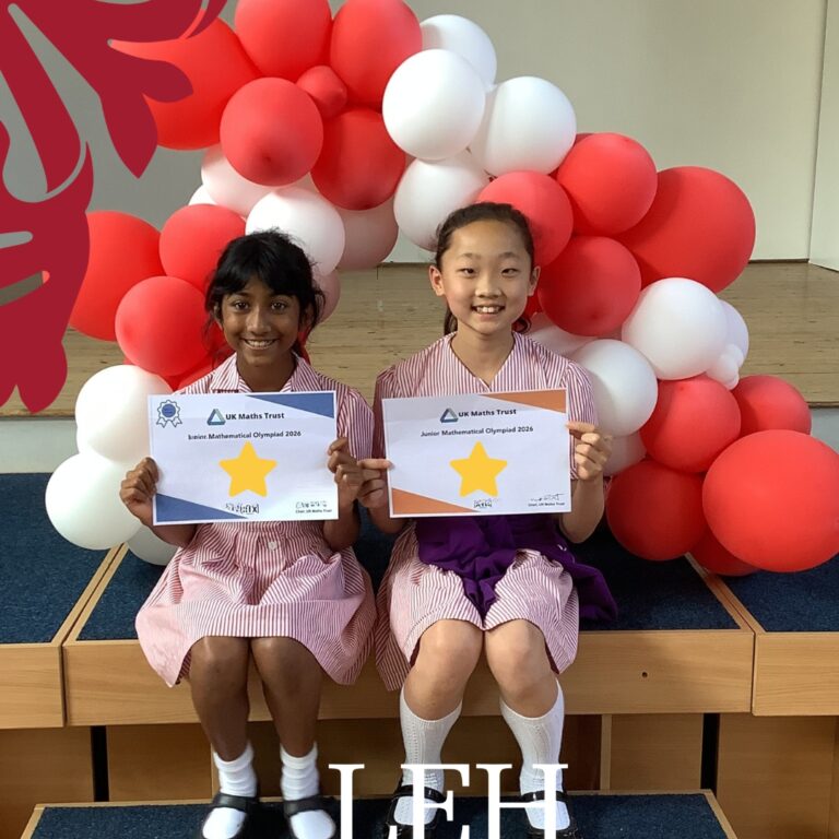 Two smiling girls in school uniforms sit on a stage holding certificates with a gold star, in front of a red-and-white balloon arch.