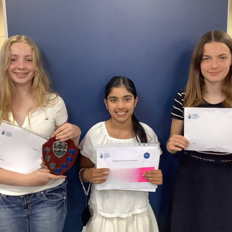 Three smiling girls stand against a blue wall holding awards: left girl in a white top with a shield trophy and certificate, center girl in a white dress holding envelopes, right girl in a striped top holding an envelope.