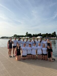 Group of young athletes in white shirts with medals posing on a lakeside dock near tents in the background.