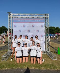 Group of young athletes in white NSR shirts posing for a photo in front of a banner and metal stage truss outdoors at a sunny event with medals around their necks.