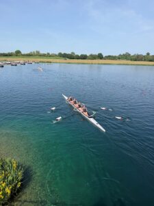 Eight rowers in a racing shell row across a calm blue lake with a docked area and grassy shoreline in the background.