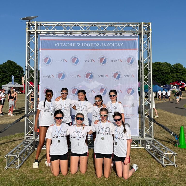Group of young athletes in white NSR shirts posing for a photo in front of a banner and metal stage truss outdoors at a sunny event with medals around their necks.