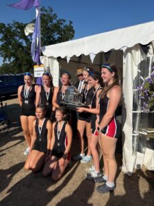 A group of young female athletes in black uniforms with medals around their necks pose outside a white tent, proudly holding a large trophy together.