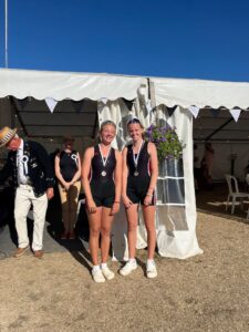 Two young female swimmers in black racing suits with medals pose in front of a white event tent, smiles on their faces as others stand nearby.