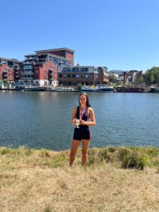 Girl in a black athletic outfit stands on a grassy riverbank, holding a trophy with a medal around her neck, with boats and red-brick buildings in the background on a sunny day.