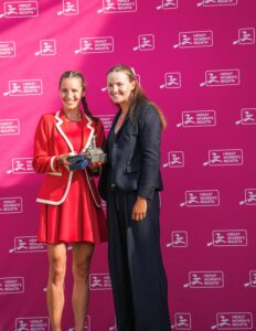 Two women pose with a trophy at Henley Women's Regatta; left in red, right in a dark blazer, against a pink backdrop with white regatta logos.