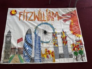 Child’s cloth banner showing a red dragon, the London Eye, Big Ben, and city buildings with colorful paper confetti on top of a maroon background.