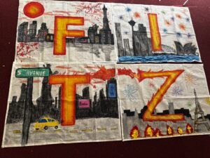 Two overlapping children’s cityscape drawings on fabric, with bright orange road shapes, dark buildings, and colorful fireworks.