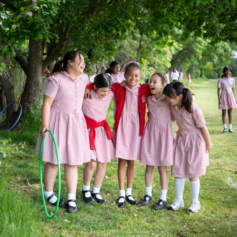 Group of schoolgirls in pink striped dresses standing together outdoors, smiling under tree shade with a hula hoop nearby.
