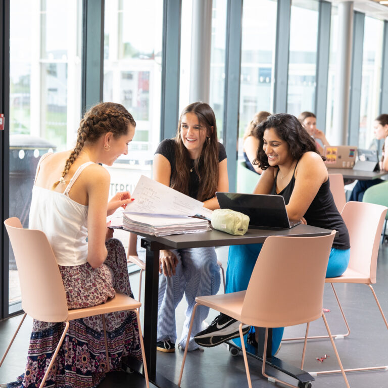 Three young women sit at a table with papers and a laptop, smiling and discussing in a bright, windowed study space.
