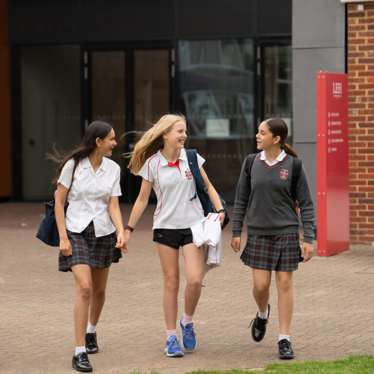 Three high school girls in plaid skirts and white shirts walk and chat on a campus walkway outside a building.