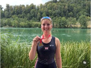 Smiling woman in a black rowing uniform by a lake, holding a gold medal on a red ribbon.
