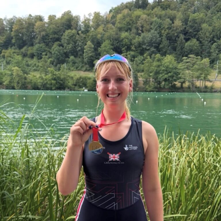 Smiling woman in a black rowing uniform by a lake, holding a gold medal on a red ribbon.