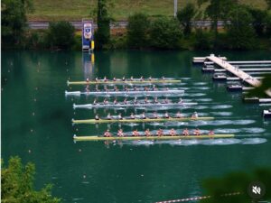 Rowers in long racing shells compete on a calm lake, parallel to each other with a dock to the right and trees in the background.
