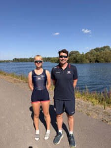 Two athletes stand by a lake on a sunny day, wearing navy athletic suits and sunglasses.