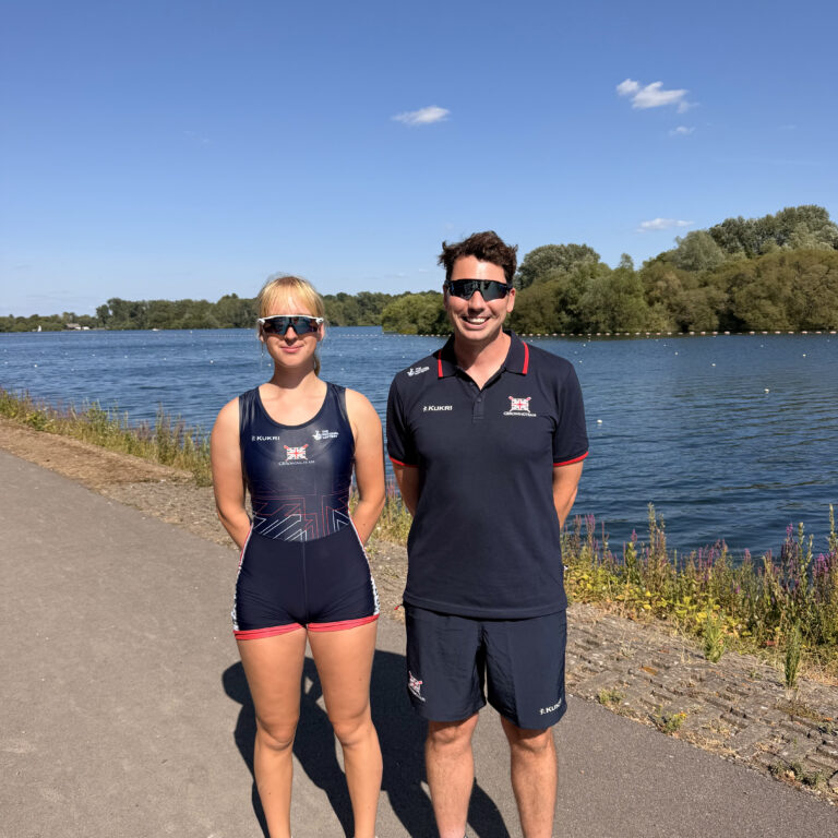 Two athletes stand by a lake on a sunny day, wearing navy athletic suits and sunglasses.