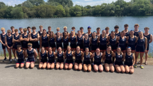 Large group of rowers in matching navy uniforms posing for a team photo by a lake with trees in the background.