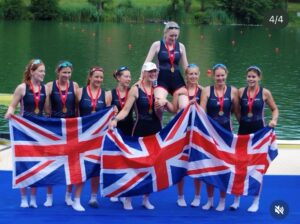 Women’s rowing team on a boat dock, wearing matching uniforms, holding British flags and wearing medals in a lake setting after a race