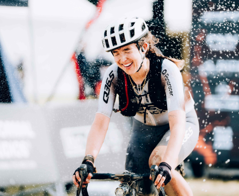 Female cyclist wearing a white helmet and race kit, smiling as water spray flies around during a race ground scene