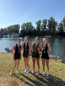 Four young female rowers in black uniforms with red stripes, wearing medals, stand by the river making peace signs after a race. Behind them are boats and a tree-lined shore on a sunny day.