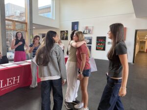 Group of students chatting and one pair hugging near a table with a red cloth in a bright gallery space with artworks on the walls.