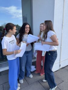 Four young women stand outside a building, smiling as they look at and share papers together.