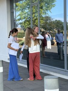Two girls hug in front of a large glass wall while another student stands nearby with papers in hand.
