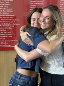 Two women smiling and hugging indoors, one in a denim jacket and the other in a light lace top with sunglasses on her head.