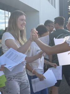 Smiling students outside a blue building give a high-five while holding papers and worksheets; casual group photo.