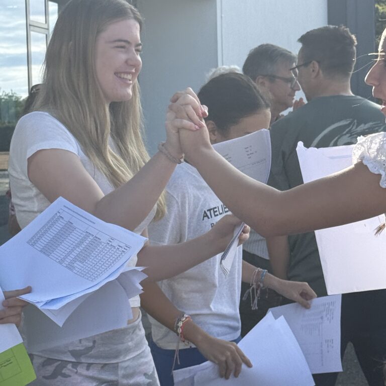 Smiling students outside a blue building give a high-five while holding papers and worksheets; casual group photo.