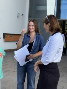 Two women stand outside chatting, one in a denim shirt holding papers and a smartphone while the other in a light top and dark skirt listens nearby.
