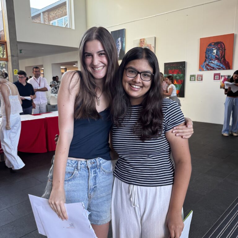 Two young women pose together in an art gallery, smiling, holding papers and bags in a bright space with artwork on the walls behind them.